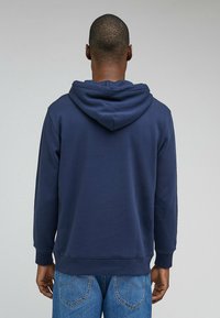Sudadera azul marino con una textura suave, que presenta una gran capucha y puños acanalados. Se lleva con jeans de mezclilla, mostrando una vista relajada desde atrás.