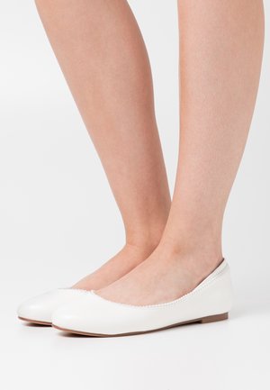 Paire de pieds portant des ballerines blanches ornées de petites perles décoratives sur les bords, sur un fond blanc uni.