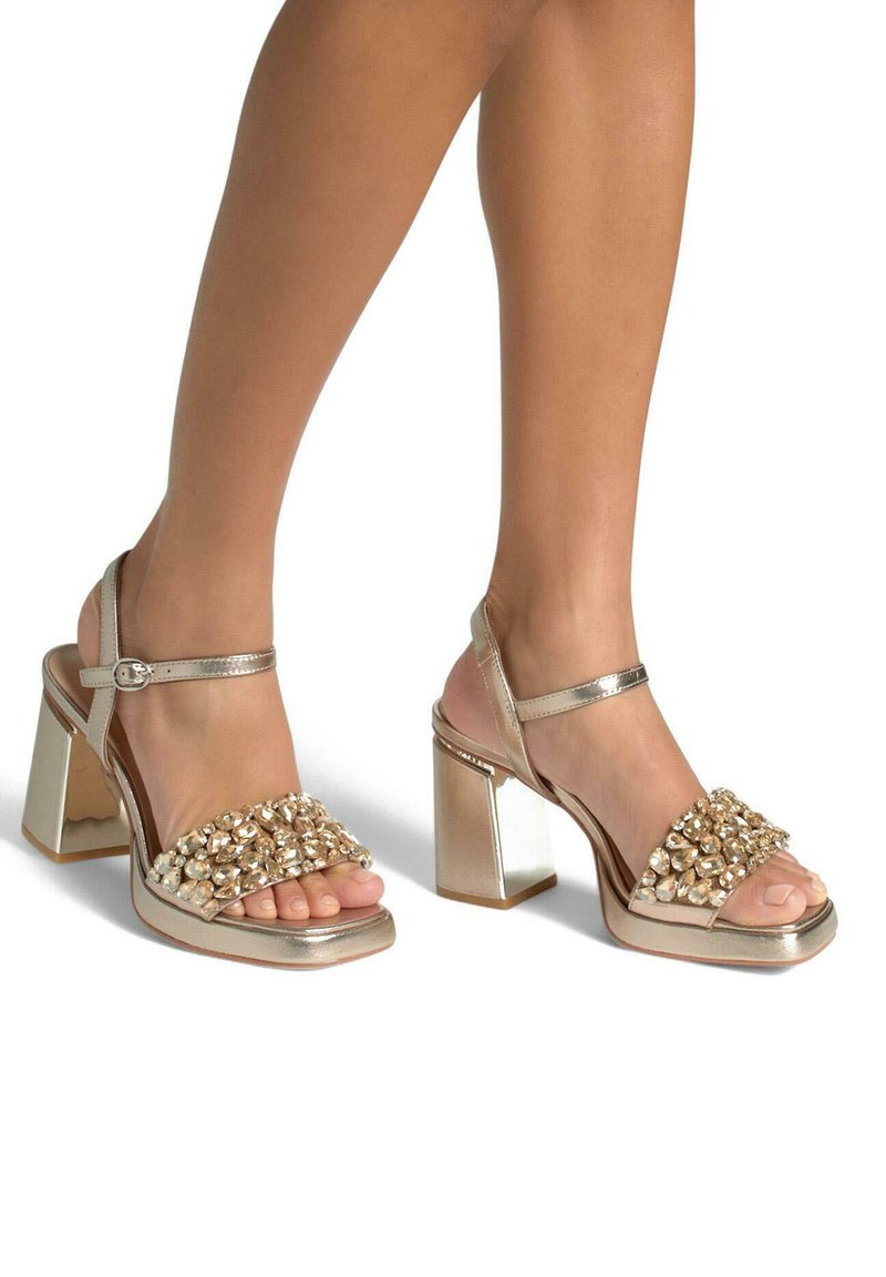 Gouden sandalen met hakken, voorzien van een brede band versierd met heldere edelstenen, een chunky blokhak en een verstelbare band rond de enkel.