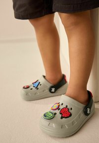 Graue Slipper mit verschiedenen bunten Charakter-Patches, strukturiertem Design und einem kontrastierenden schwarzen Fersenriemen, getragen von einem Kind.