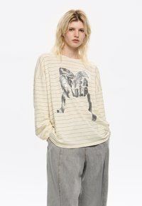 Camicia a maniche lunghe a righe giallo chiaro con una grafica metallica di un fiocco e testo sul davanti, abbinata a pantaloni larghi grigi.