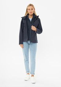 Marine waterdichte jas met capuchon, ritsluiting en trekkoordaccenten, gedragen over een gewatteerd vest. Lichte jeans en witte sneakers maken de look compleet.