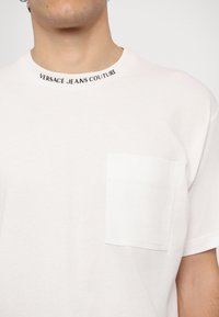 Balts kokvilnas T-krekls ar apaļu kakla izgriezumu, ar melnu uzrakstu "VERSACE JEANS COUTURE" uz kakla un kreisās krūtis kabatu.