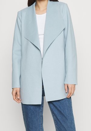 Vrouw draagt een lichtblauwe openvallende jas over een wit shirt en een blauwe spijkerbroek, staand tegen een effen achtergrond.