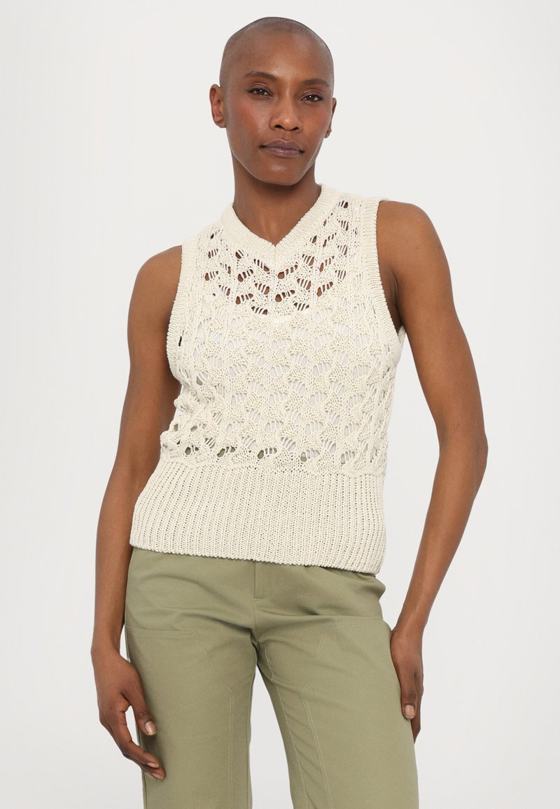 Haut en tricot sans manches crème avec un motif en dentelle sur le corsage et un ourlet à côtes, associé à un pantalon vert. Texture lisse, design en V.