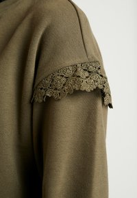 Sudadera de color verde oliva con un delicado encaje en los hombros, destacando intrincados diseños florales y una superficie de tela suave.