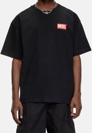 Personne portant un T-shirt noir oversize avec un patch logo Diesel rouge sur la poitrine gauche et un collier chaîne argenté.