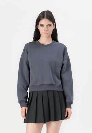 Grå, kort sweatshirt med lange ermer, rund halslinning og ribbestrikkede mansjetter. Kombinert med en mørk, plissert skjørt. Enkel, minimalistisk design.