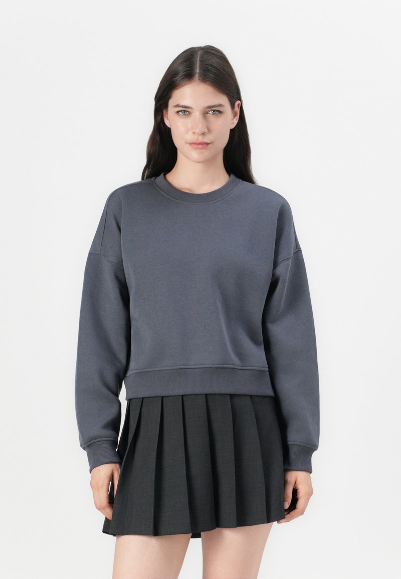 Sweatshirt cinza, cropped, com mangas longas, decote redondo e punhos canelados. Combinado com uma saia pregueada escura. Design simples e minimalista.
