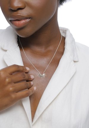 Gros plan sur une femme portant un collier soleil argenté, le touchant délicatement, vêtue d'un blazer blanc à décolleté plongeant.