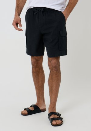 Mand iført sorte cargoshorts med sidelommer og sorte sandaler med spænder, stående mod en ensfarvet baggrund.