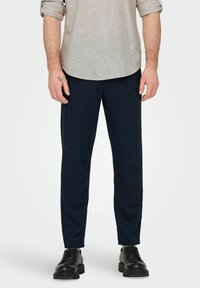 Pantaloni blu navy realizzati in tessuto leggero, con una vestibilità dritta, un leggero lucido e tasche laterali. Indossati con una camicia di colore chiaro.