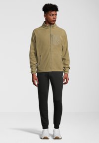 Giacca in pile con zip di colore verde oliva, colletto alto, tasca frontale e zip laterali, abbinata a pantaloni della tuta neri e sneakers bianche.