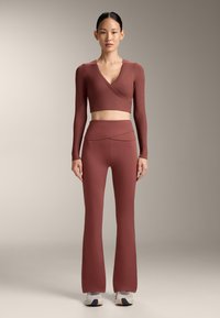 Modella in piedi, indossa un top incrociato corto a maniche lunghe color bordeaux abbinato a pantaloni a zampa vita alta e sneakers bianche.