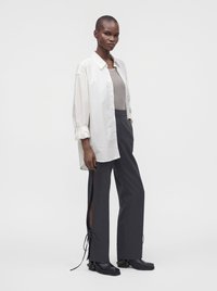 Personne grande portant une longue chemise blanche boutonnée, un haut beige, un pantalon large foncé avec des fentes latérales noué aux chevilles, et des chaussures noires, debout sur un fond blanc.