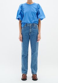 Blå t-shirt med puffärmar som matchas med högmidjade ljusblå denimjeans. Modellen har på sig bruna vävda skor. En enkel vit bakgrund.