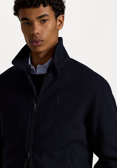 Giacca di lana blu navy con zip frontale e colletto alto, caratterizzata da una texture sottile. Indossata sopra una camicia a righe con colletto.