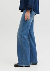 Sinised teksapüksid, mis on alt laieneva lõike ja keskmise taljega, näitamaks sirget lõiget ja kerget pleekimist. Kandmisel mustade kingade ja tumeda ülaosaga.