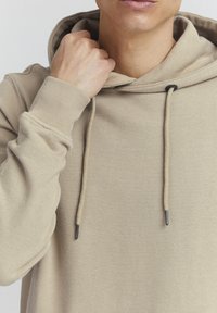 Bež hoodie od mekog materijala, s kapuljačom na vezanje, rebrastim manžetama i teksturiranom završnicom. Približavanje na izrez i ruku.