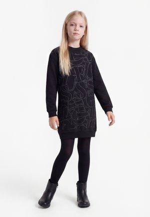 Jeune fille aux longs cheveux blonds portant une robe sweat noire avec des lettres blanches en pointillé, des collants noirs et des bottines noires.