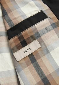 Tissu à carreaux avec des rayures beige, marron et noir et une petite étiquette rectangulaire blanche cousue affichant le nom de la marque NEXT.