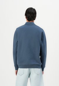 Mørkeblå genser, lange ermer, ribbestrikket krage og mansjetter, mykt stoff, avslappet passform. Kombinert med lyseblå jeans, vises bakfra.