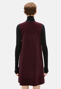 Burgunderfarbendes Cordkleid mit ärmellosem Design, kombiniert mit einem schwarzen Langarmrollkragenpullover darunter. Glatte Textur, einfarbig.