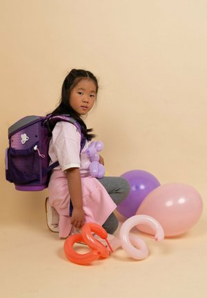 Lila Rucksack mit einem Netzfach, graue Leggings, rosafarbene Ballons und ein lila Ballontier. Heller Hintergrund. Ein Kind hockt zwischen den Gegenständen.