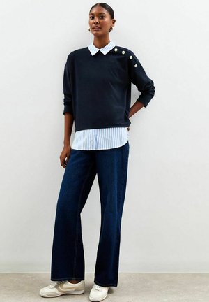 Mujer vestida con un suéter azul marino con botones dorados en el hombro, camisa a rayas azul y blanca, jeans de pierna ancha y zapatillas beiges, de pie en el interior.