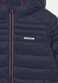 Navy blue puffer jacket with horizontal quilted sections, red and white zipper, and a small rectangular logo patch on the chest.