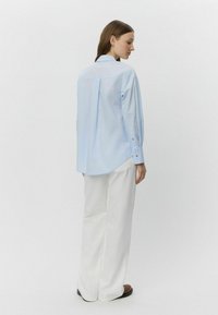 Cămașă lungă de culoare albastră deschisă cu detaliu la spate, manșete cu butoane și o croială relaxată, asortată cu pantaloni largi albi și pantofi maro.