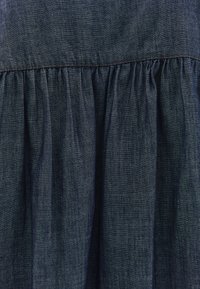 Denimová šaty s tmavě modrou tkaninou a nabíranou sukní, viditelným šitím podél švu a hladkým, mírně texturovaným povrchem.