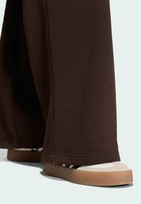 Hnedé nohavice širokého strihu s hladkou textúrou, skombinované so svetlými topánkami s gumovou podrážkou a čiernymi pruhmi na vrchu.