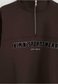 Sudadera marrón con cuello alto, con cierre de media cremallera y texto bordado "AIM'N SPORTSWEAR EST. 2013" en blanco y negro.
