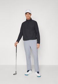 Sudadera negra con cremallera, pantalones gris claro y zapatos de golf blancos. El conjunto incluye una gorra blanca y un palo de golf en la mano del modelo.