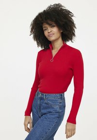 Rotes, geripptes Langarmtop mit halbem Reißverschluss und rundem Zug, kombiniert mit hochgeschnittenen blauen Jeans. Die Textur ist glatt und enganliegend.
