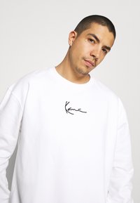 Witte sweatshirt met lange mouwen en een zwart handtekeninglogo op de borst. Gemaakt van een zachte, gladde stof, met een relaxte pasvorm.