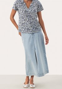 Mujer vistiendo una blusa corta de manga azul con flores, una falda midi de mezclilla azul clara con abertura frontal, y bailarinas plateadas, de pie contra un fondo blanco.