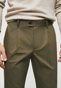 Pantaloni su misura verde oliva realizzati in tessuto liscio, con piega frontale, passanti per cintura e chiusura a un bottone in vita.