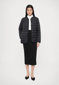 Kvinne står mot hvit bakgrunn iført svart quiltet jakke, hvit høyt halsplagg, svart midi-skjørt og svarte spisse sko.