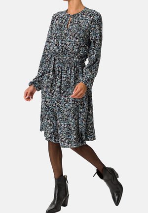 Femme portant une robe fleurie mi-longue à manches longues avec un lien à la taille, des collants noirs transparents et des bottines noires à talons.
