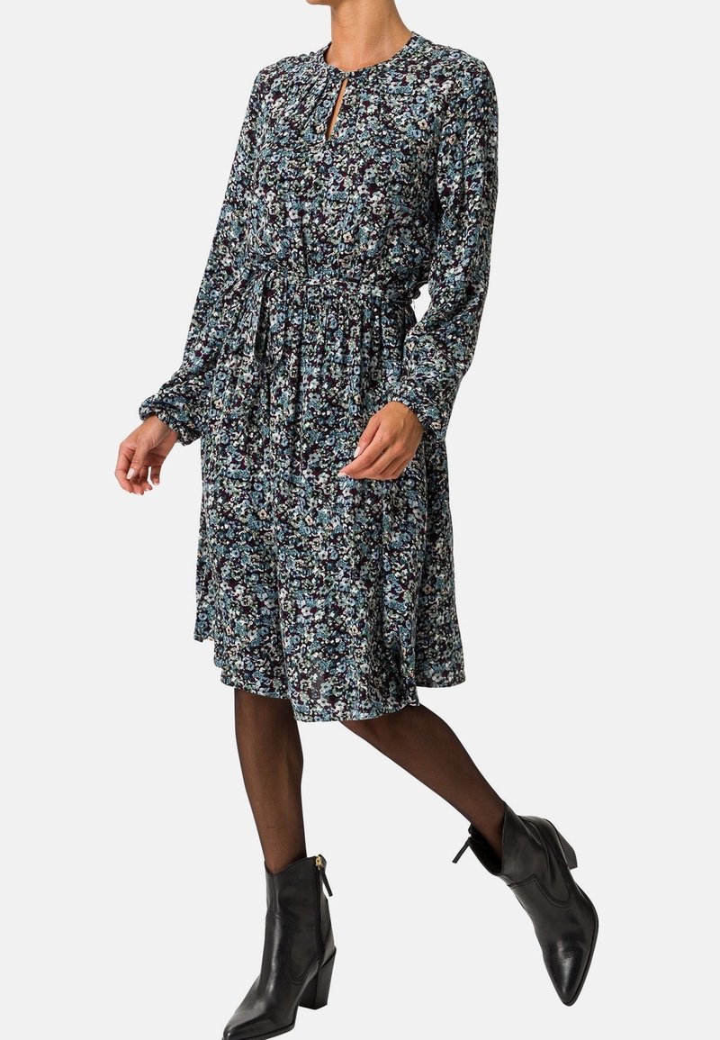 Femme portant une robe fleurie mi-longue à manches longues avec un lien à la taille, des collants noirs transparents et des bottines noires à talons.