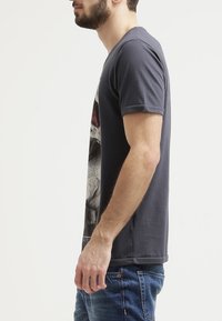 Šedé bavlněné tričko s krátkým rukávem, s grafickým potiskem na přední straně a rovnym lemem. Nosené s modrými džínami.