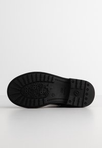 Semelle de chaussure noire avec des profondes rainures, des indentations circulaires et en forme de croix, et un numéro de taille visible "29" sur un fond blanc.