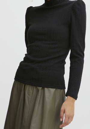 Strak zittend zwart textuur lang mouw shirt met pofschouders, gecombineerd met hooggetailleerde, olijfgroene wijde broekspijpen, gedragen door een staande persoon.