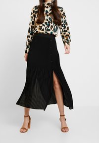 Mustat pleat-hameet, joissa on etureikä ja nappidetaileja. Yhdistetty kuviolliseen pitkäharmaaseen puseroon ja vaaleanruskeisiin korkokengiin. Suuri tekstuuri.