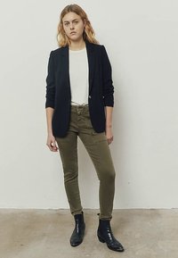 Zwarte blazer, wit T-shirt, olijfgroene slim-fit broek en zwarte enkellaarsjes met studs. Eenvoudig ontwerp, geseald blazer en gestructureerde broek.