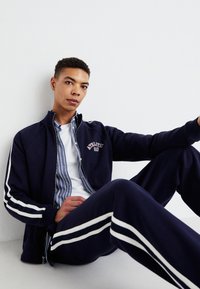 Marine tracksuit med hvite striper, jakke med glidelås som har teksten "Athletic", lagd over en skjorte med blå striper.