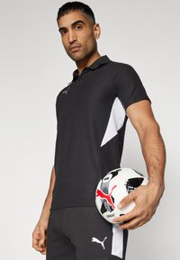 Polo noir avec des accents blancs sur les côtés, manches courtes et col. Tient un ballon de football noir et blanc avec des accents rouges.