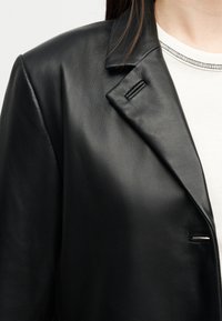 Μαύρο πέτο δερμάτινου τζάκετ πάνω από λευκό τοπ που φοριέται από άτομο με σκούρα μαλλιά ορατά στον ώμο.